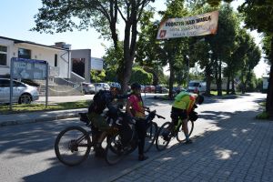 Trójka rowerzystów pozujących do zdjęcia pod banerem powitalnym, zawieszonym nad drogą