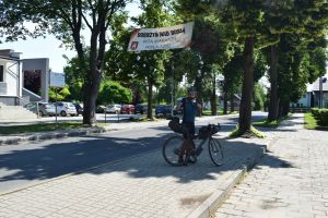 Rowerzysta pozujący przy rowerze do zdjęcia, nad nim rozciągnięty duży baner powitalny
