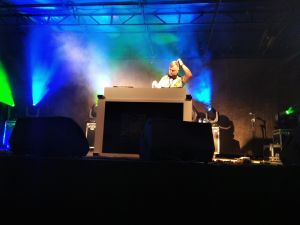 Dj stojący za konsoletą na scenie, za nim kolorowe światła-element scenografii