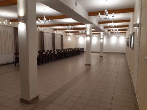 Duża sala, ściany pokryte białymi płytkami elewacyjnymi, pod sufitem podwieszone eleganckie żyrandole, na sali długi stół i...