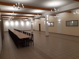 Duża sala, ściany pokryte białymi płytkami elewacyjnymi, pod sufitem podwieszone eleganckie żyrandole, na sali długi stół i...