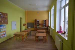Sala lekcyjna w kolorze ścian zielono-żółtym wraz z wyposażeniem-ławki, krzesła, szafki, na głównej ścianie telewizor