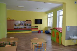 Duża sala lekcyjna, przy jednej ścianie ławki i krzesła, na drugiej szafki z zabawkami, w rogu duży domek do zabawy